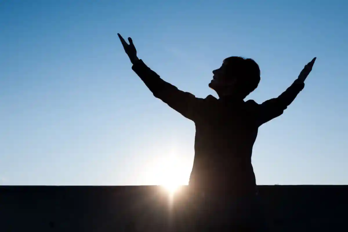 black silhouette of woman looking up and raising hands with a blue sky background
