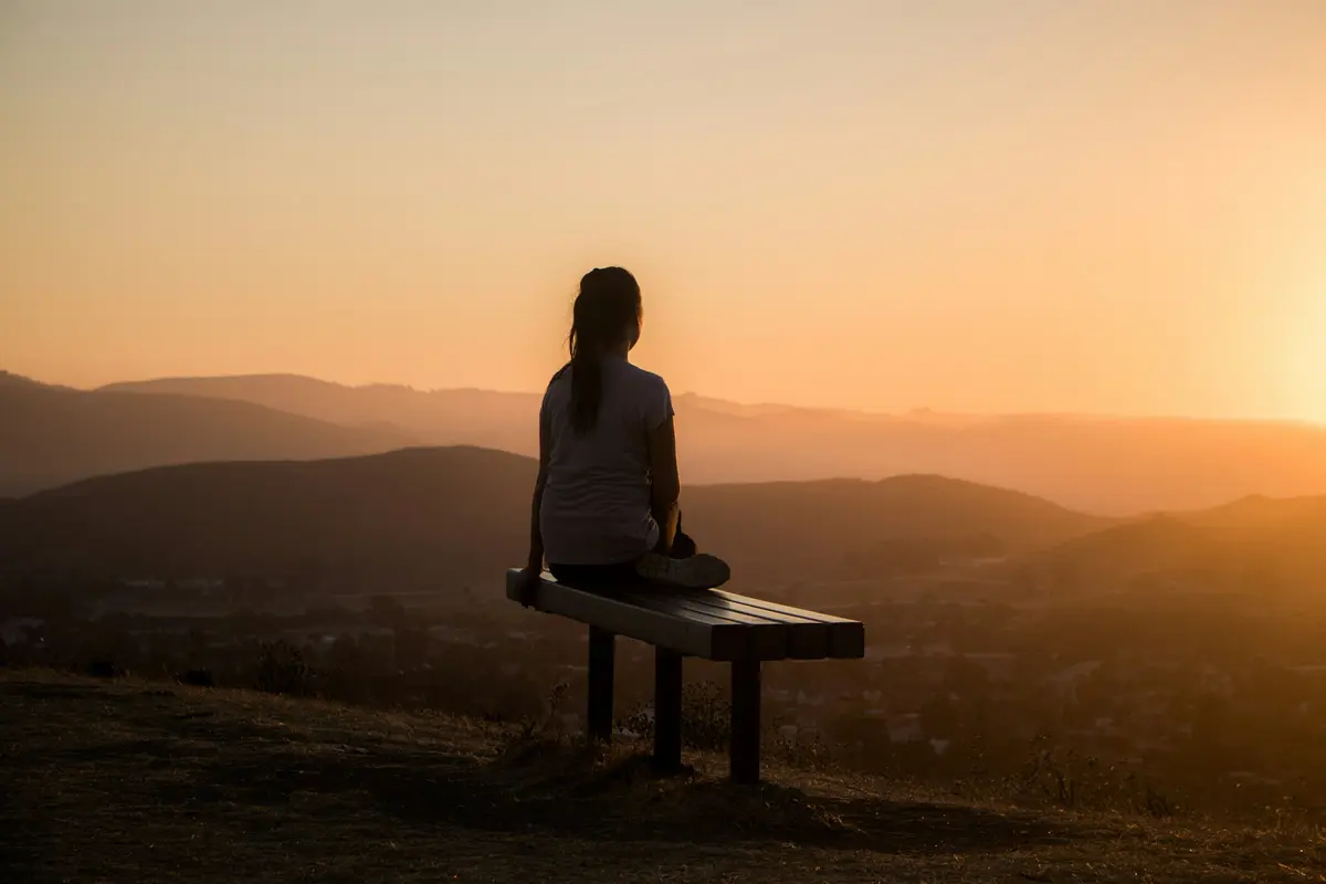 Woman sitting alone on a wooden bench looking out over hills at sunset