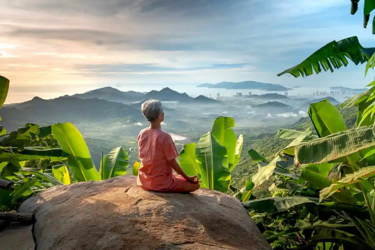 Woman meditating on a large rock overlooking a jungle, with banana plant leaves surrounding her.