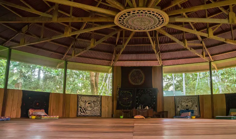 The empty maloca: a polygonal wooden ceremony hall with rafters radiating from a carved ceiling medallion, half-walls open to the forest on every side, and floor mats laid around the perimeter.