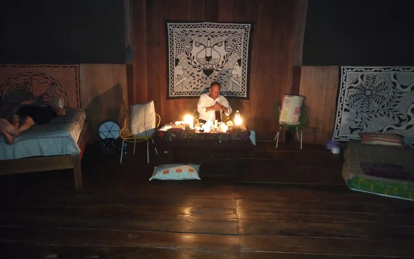 At night, a facilitator in a white shirt sits at a small altar table lit by candles beneath a jaguar-motif tapestry.