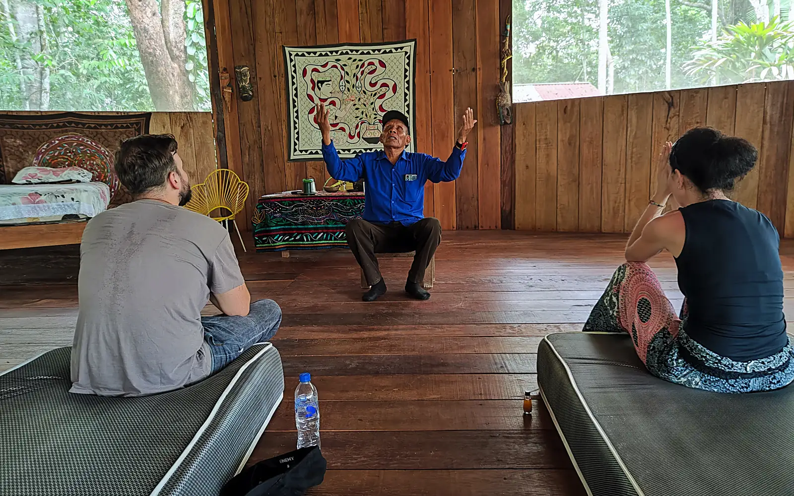 Inside the maloca, the curandero sits with both hands raised in blessing while two guests sit facing him on mats, a Shipibo textile hanging on the wall behind.