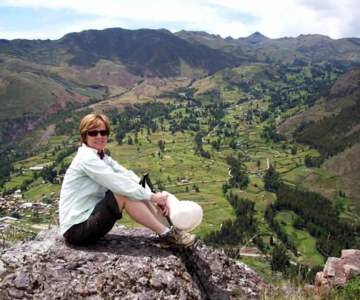 Woman in sunglasses and hiking gear sits on a rock overlooking a vast green mountain valley.