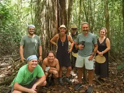 Seven smiling people, four men and three women, pose in a dense jungle by a large tree with roots