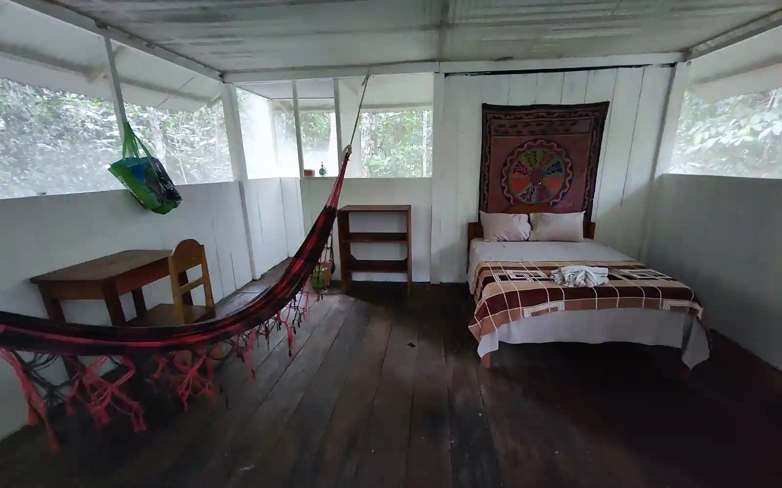 A guest room with a single bed under a woven wall hanging, a red hammock strung across the room, a desk and chair, and screened windows looking into jungle.
