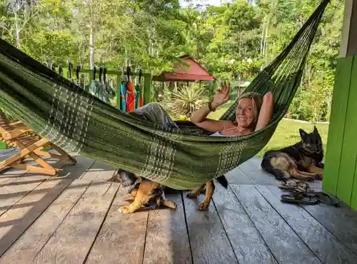 Blonde woman smiles and waves from a green hammock on a wooden porch, two dogs near her, lush jungle background