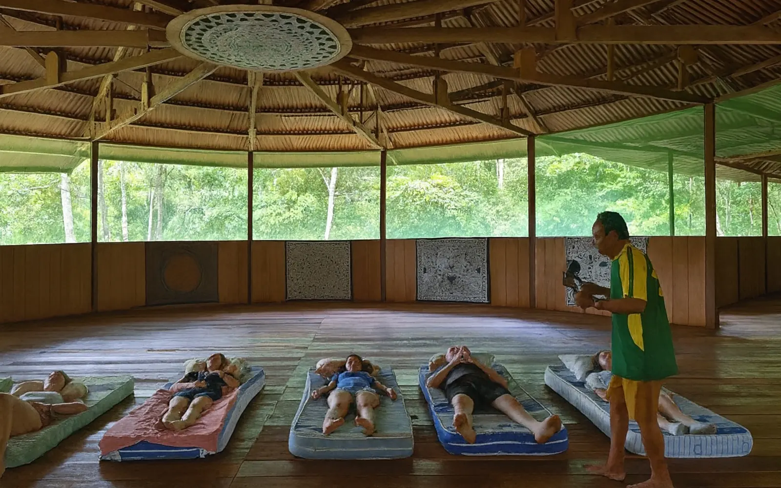 A healing round in the maloca: guests lie resting on mats across the wooden floor while the healer moves among them with a rattle, jungle visible through the open screened walls.