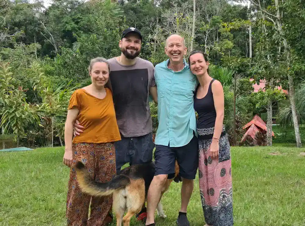 Four smiling adults and a dog stand in lush green nature; a man wears a turquoise shirt, women wear patterned pants.