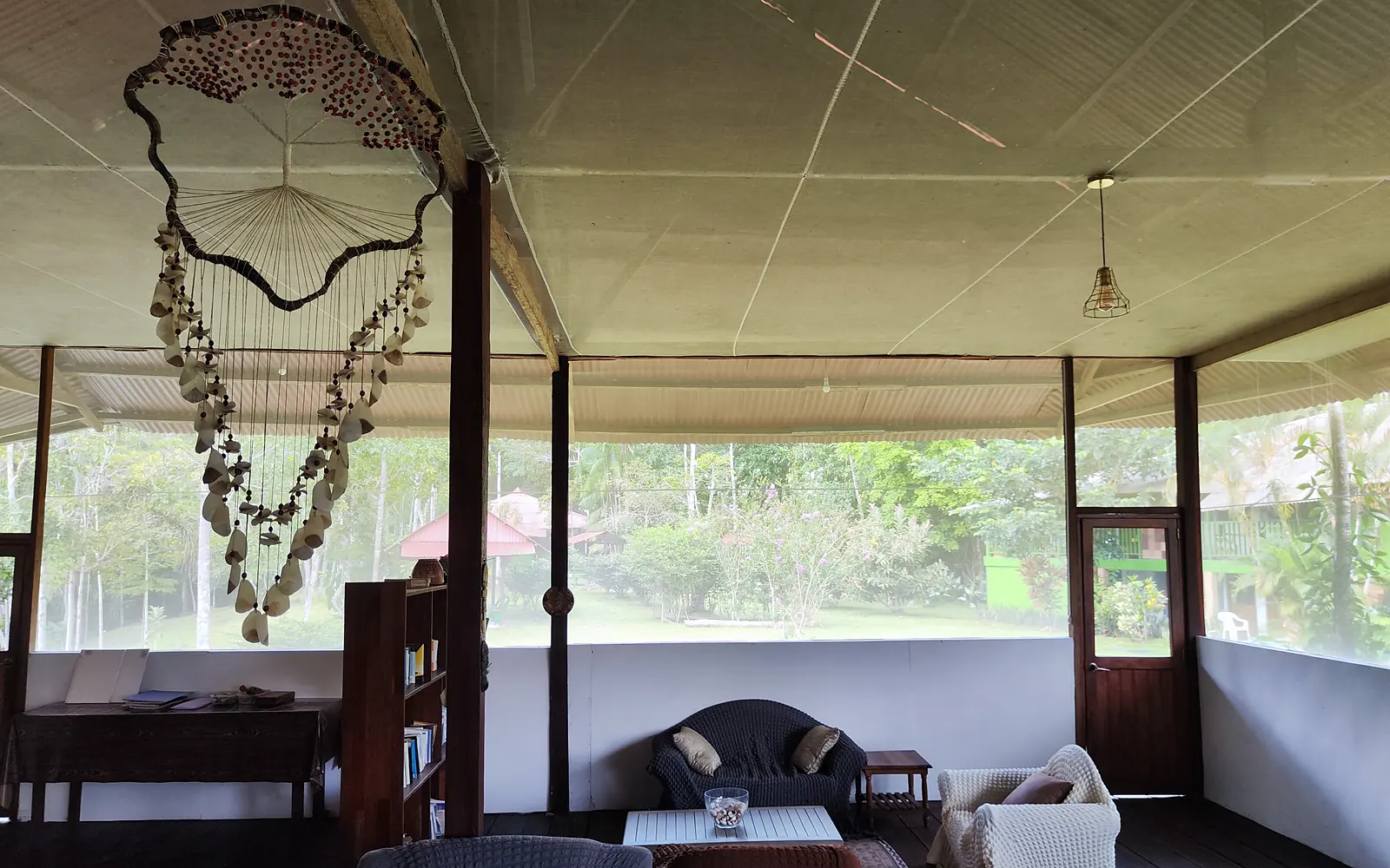 The open-air lounge: sofas and armchairs on a dark wooden floor, a large hanging ornament of shells and beads, jungle visible through the screened walls.