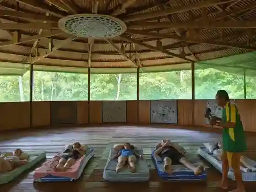 A group rests on mattresses in a round, open-air lodge with a thatched roof, surrounded by lush forest, led by a guide.