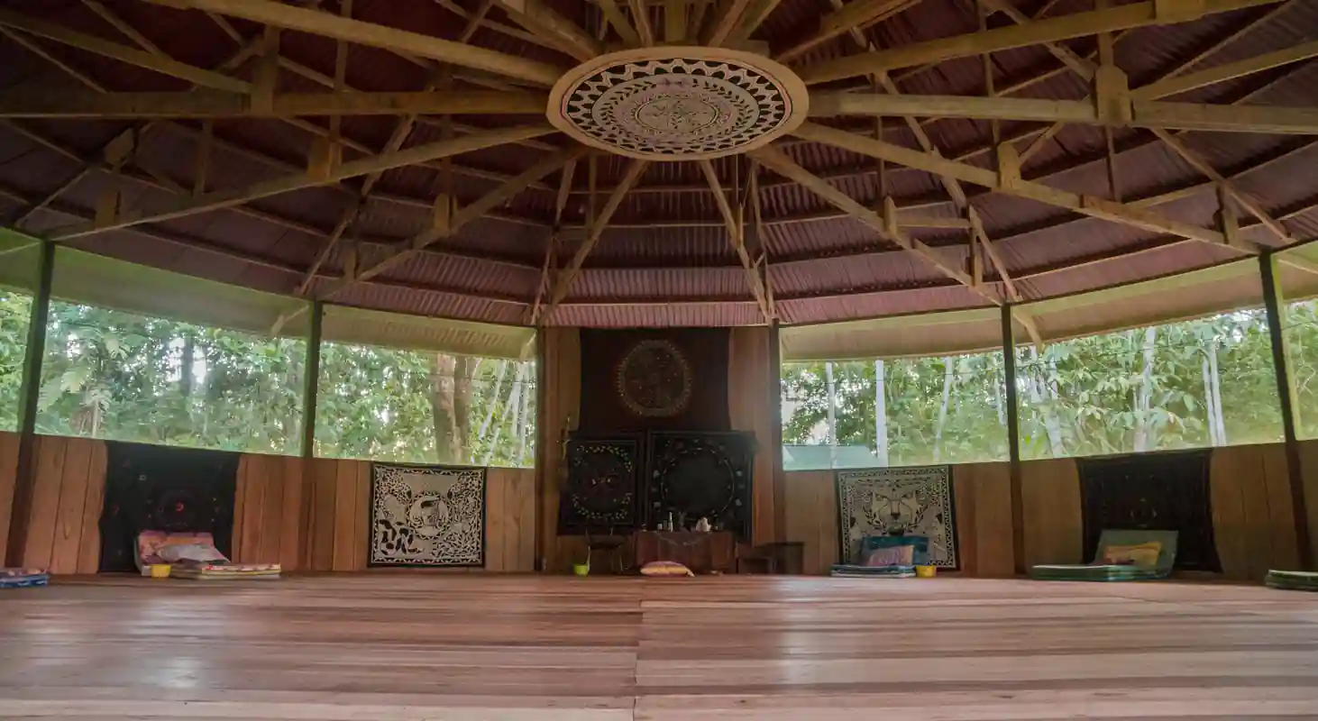 Inside of a moloca ready for an ayahuasca ceremony.  Wooden plank floor, shipibo cloths on walls, view of trees through large screened windows