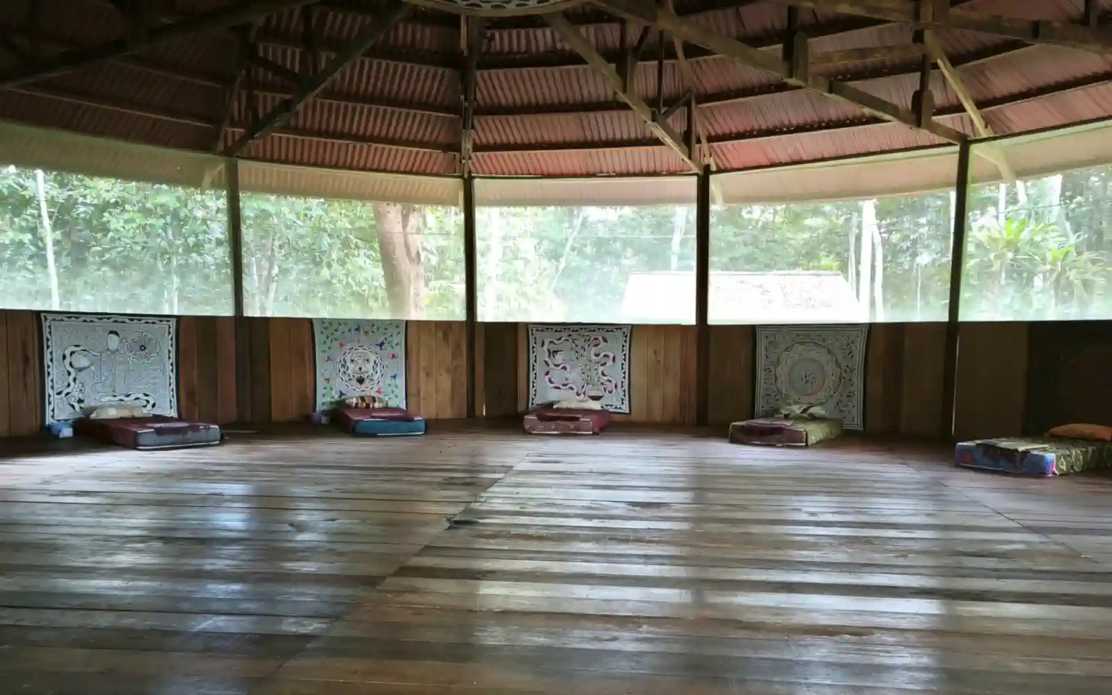 Wide interior view of the round maloca with five sleeping mats set out along the curved wall, each beneath its own patterned cloth, jungle visible through the screens.