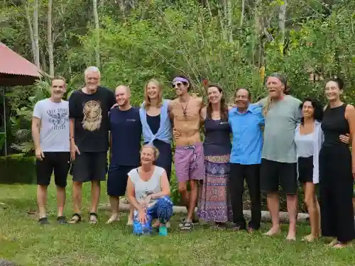 A group of 12 people, mostly standing, pose outdoors on grass with dense green foliage behind them