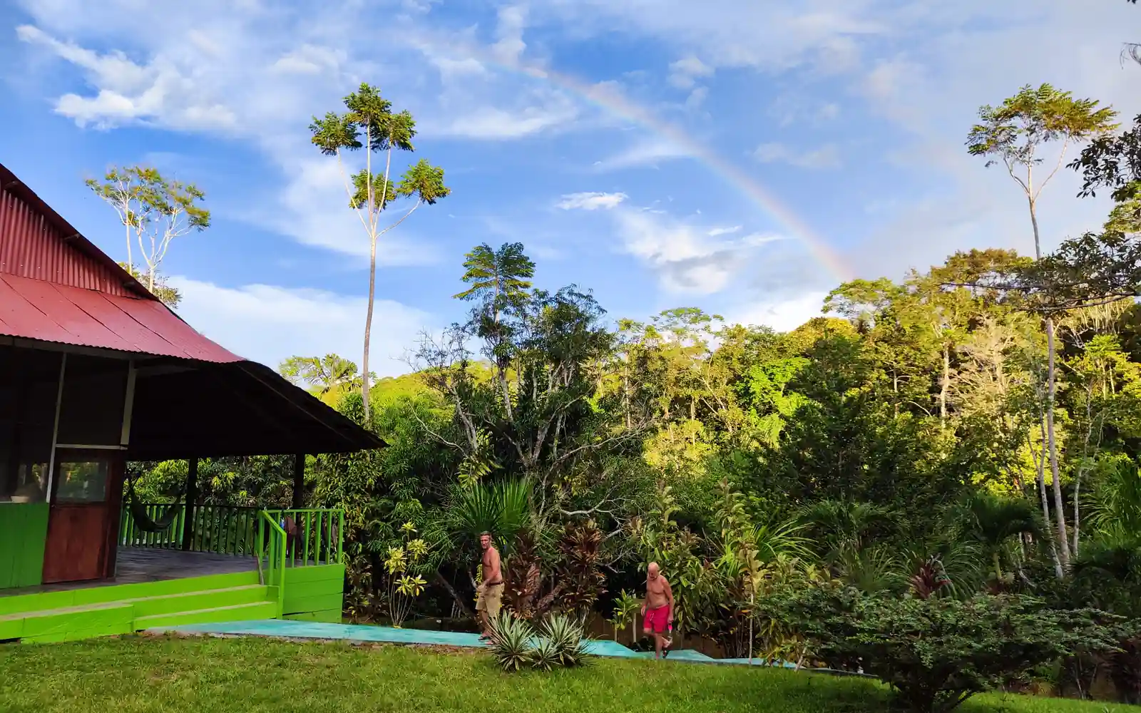 A red-roofed house with green trim next to a lush jungle under a blue sky with a faint rainbow Two people walk on a path.