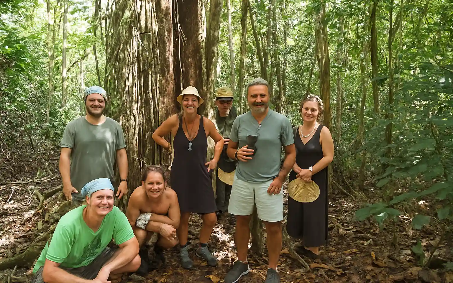Seven smiling people, four men and three women, pose in a dense jungle by a large tree with roots