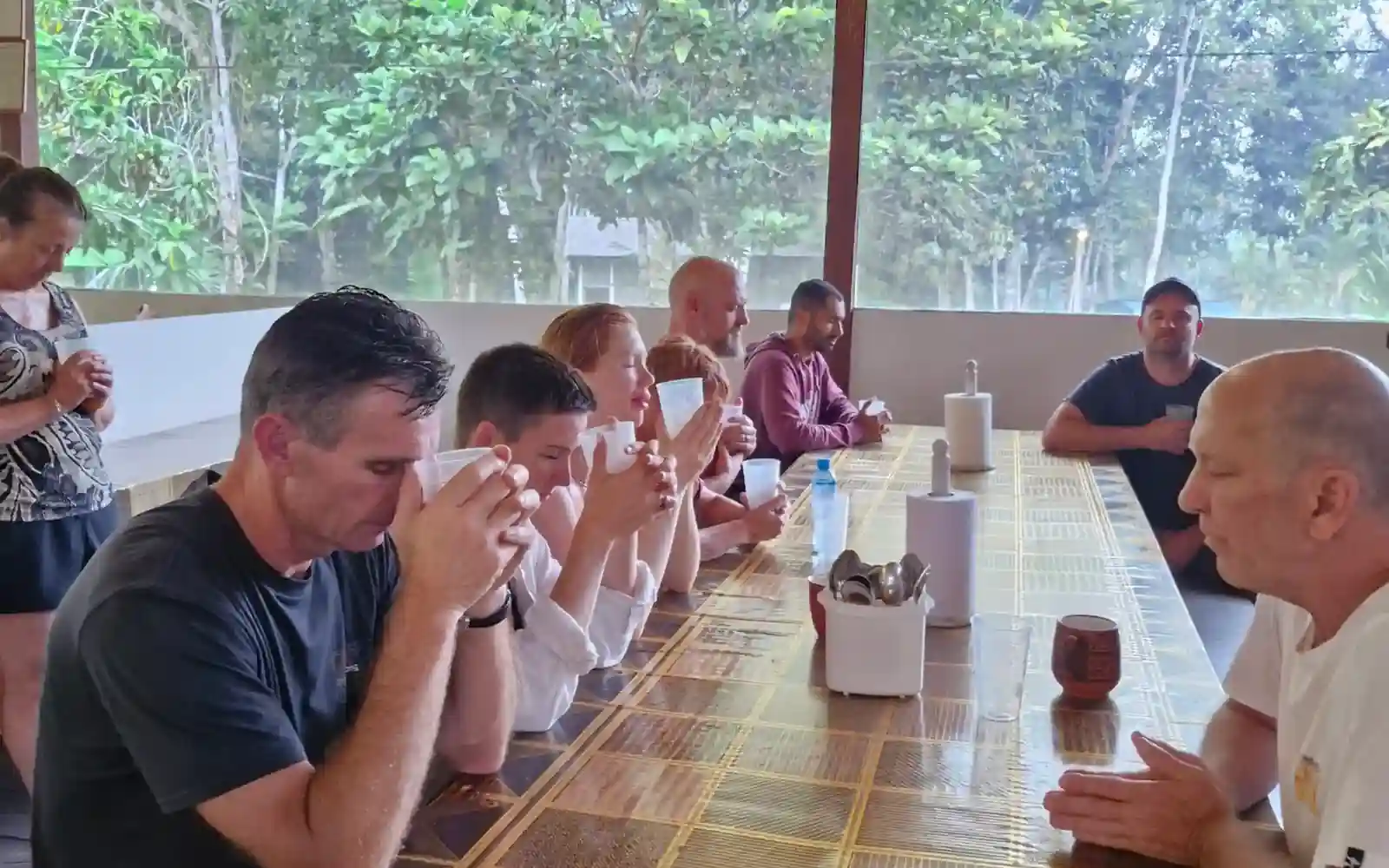 People gathered at a long patterned table, one man sips from a white mug, another gestures, with large windows showing lush greenery
