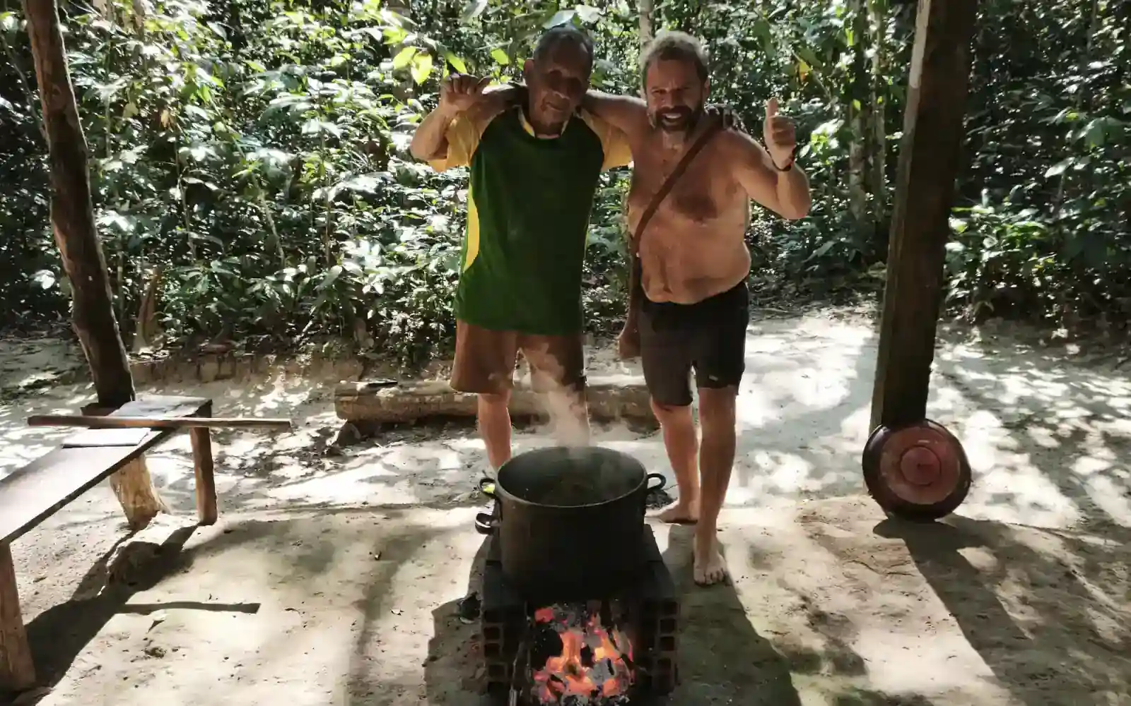 Two men stand behind a large pot steaming over an open wood fire in a sandy clearing among jungle trees, one in a green and yellow shirt, the other giving a thumbs-up.