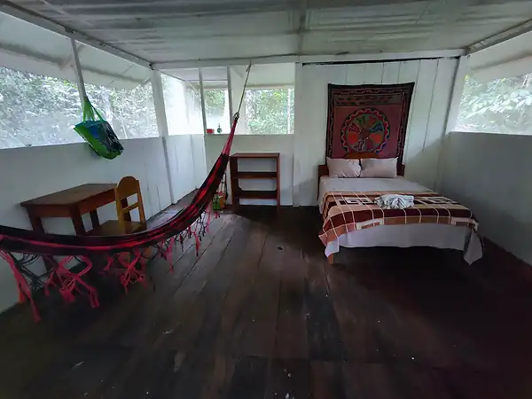 A guest room with a single bed, a woven wall hanging, a red hammock strung across the room and a small desk.