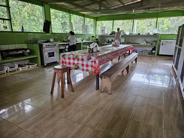 The open sided kitchen and dining hall with a long table, benches and two kitchen staff at work.