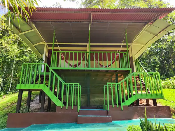 A two story guest cabin on stilts with green railings, twin staircases and hammocks strung on the upper porch.