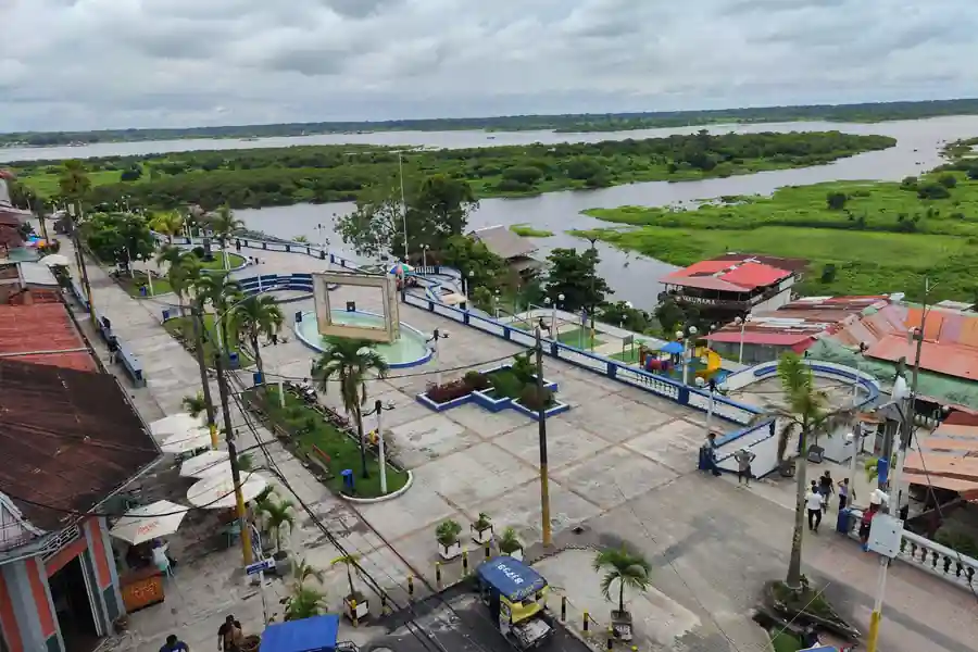 An aerial view of a riverside plaza with a circular fountain and palm trees, overlooking the wide river beyond.