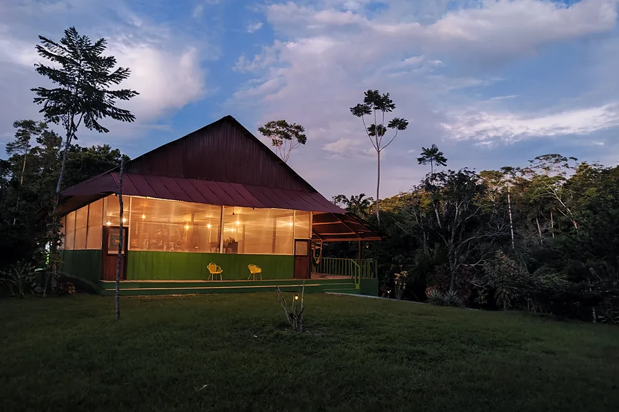 The lodge at dusk, its screened walls glowing with warm interior light beneath a purple sky, jungle dark on either side.