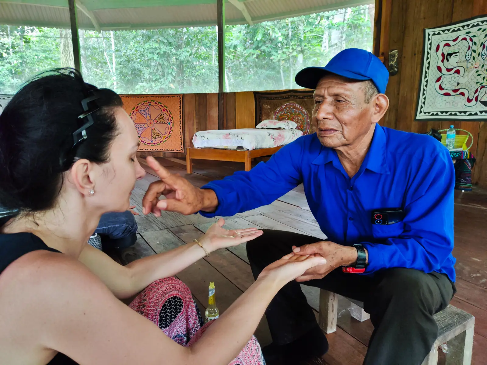 Man in blue cap points at woman's nose, holding her hands in a wooden room with cultural tapestries.