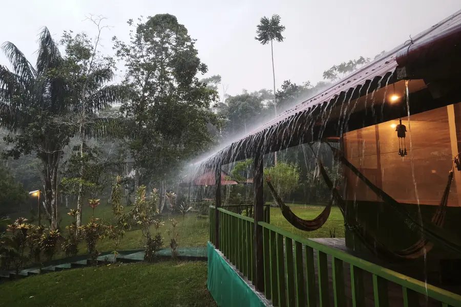 Heavy rain pouring in a sheet off the maloca roof, hammocks hung along the open side, jungle grey with rain beyond