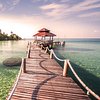 Profile picture Curved wooden pier leading to a thatched gazebo over clear turquoise water, with a tropical coast in the distance.